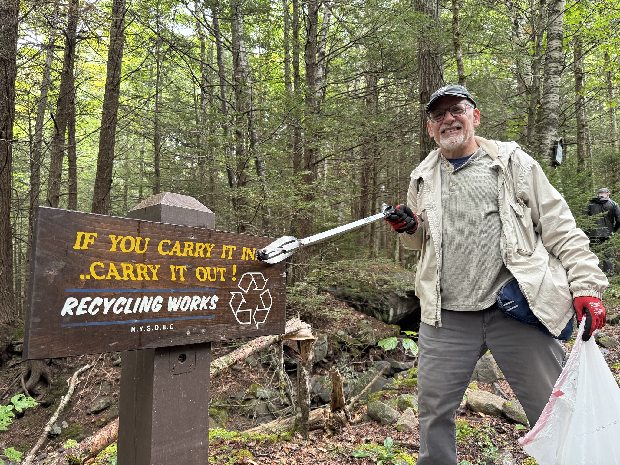 A man holding a trash bag smiles next to a sign reading 'If you carry it in, carry it out.'