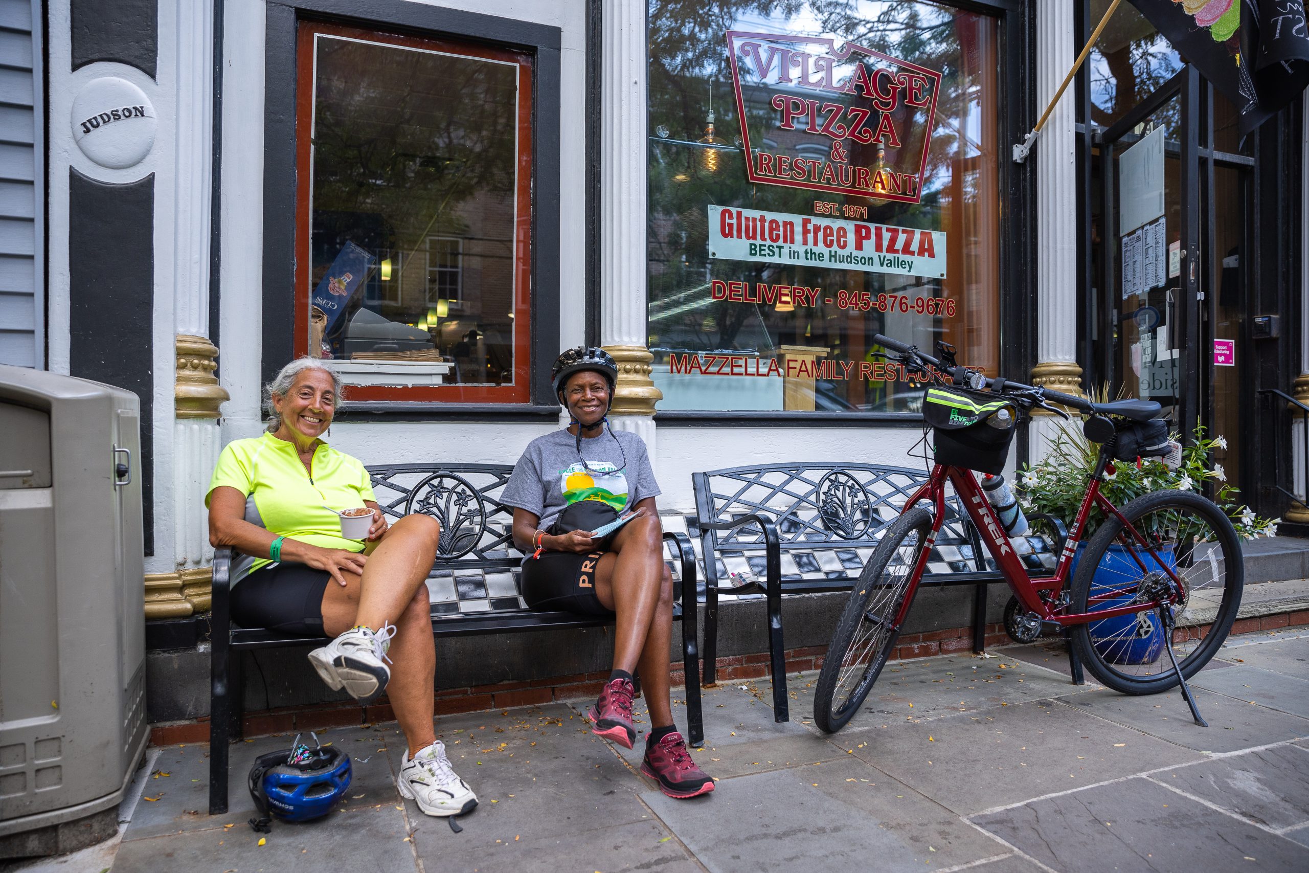 Two women in cycling gear sit outside of a pizza shop.