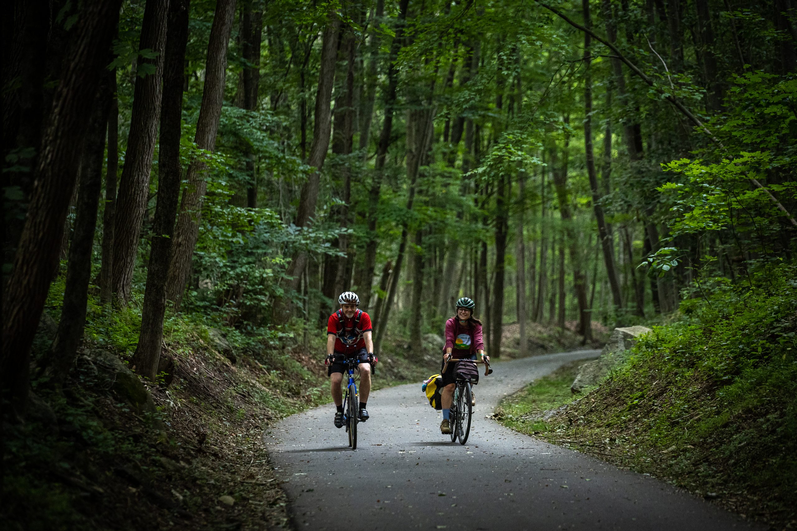 A man and a women cycle through a shaded, wooded area.