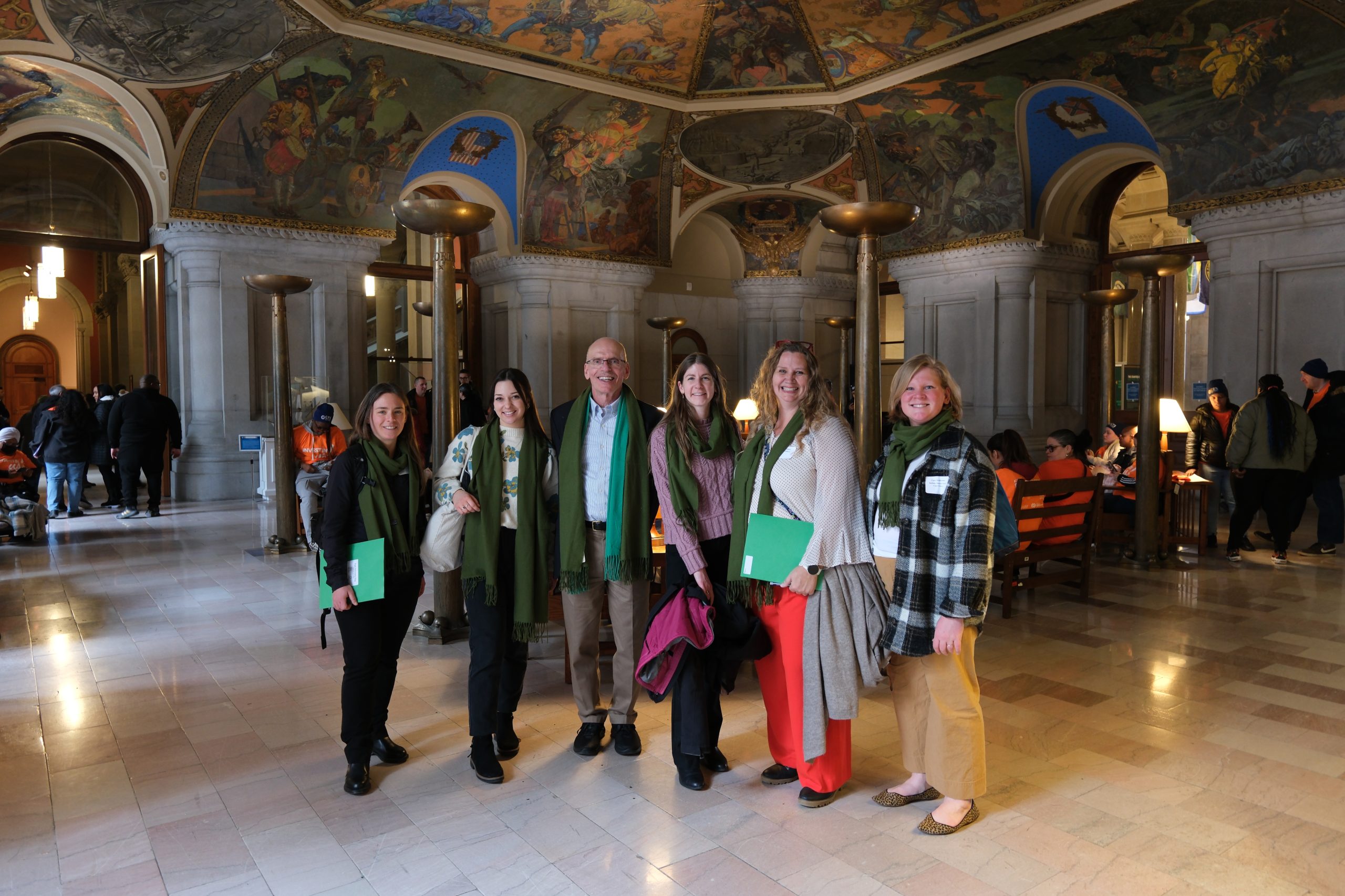 A group of advocates wearing green scarves pose for a photo at the NY State Capitol.