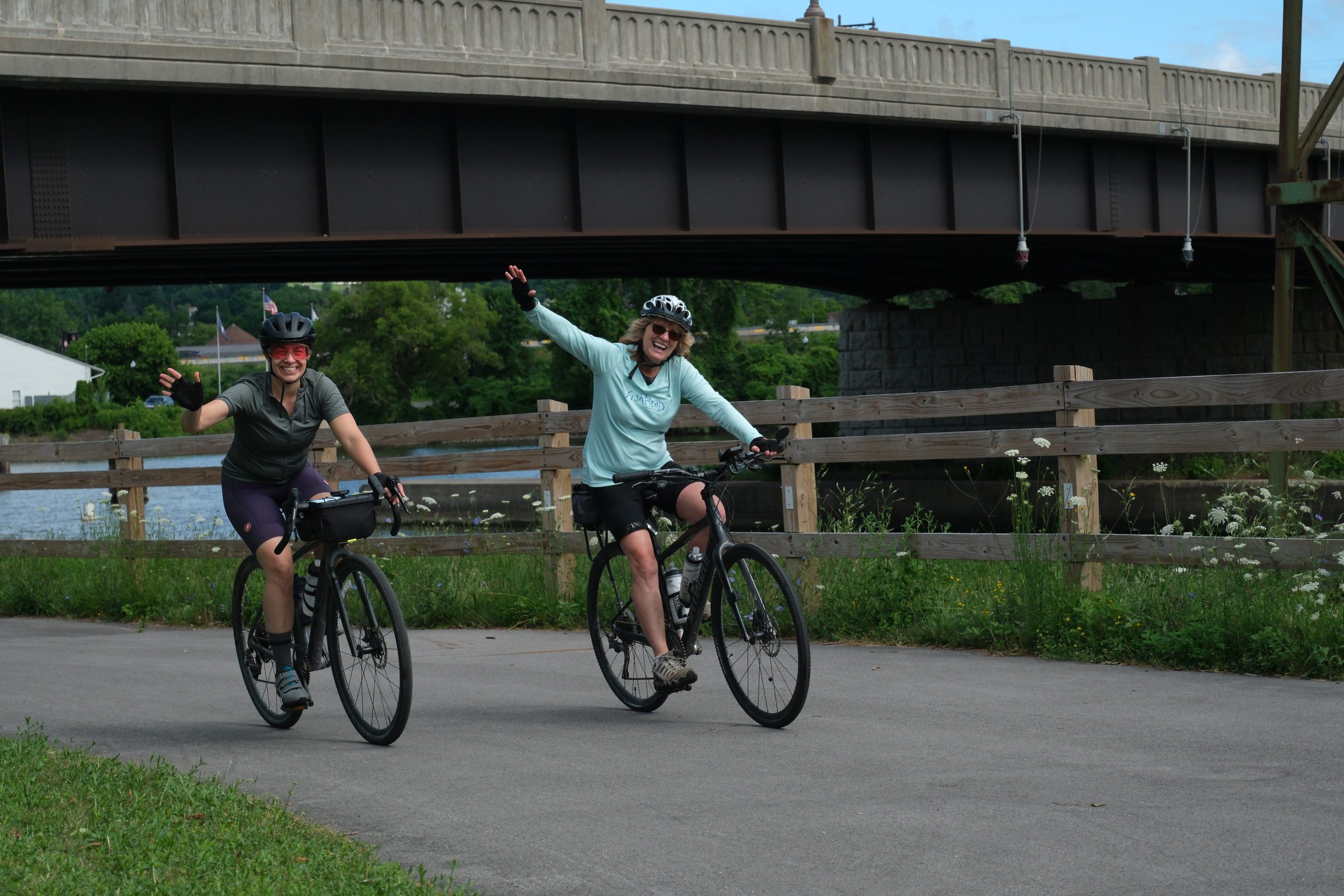 Two women smile and wave as they cycle along the Erie Canal.
