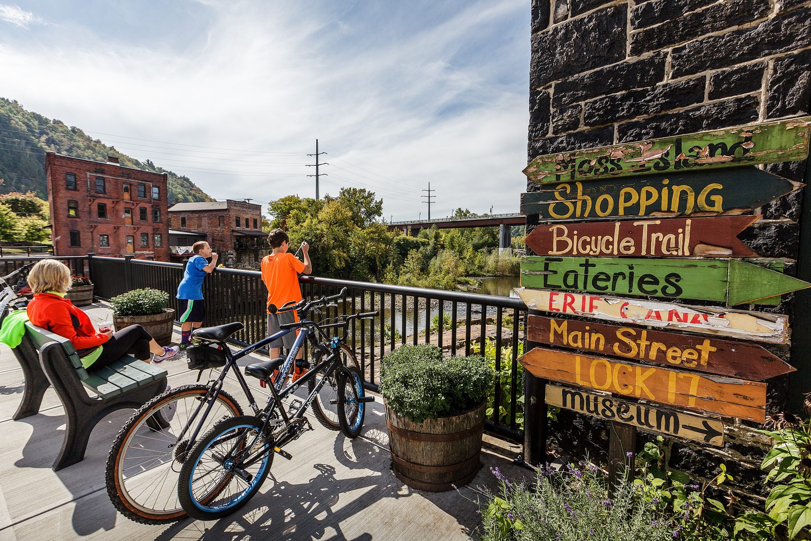 Two children stand at a railing alongside a bike and directional signage, looking down onto the Mohawk River.