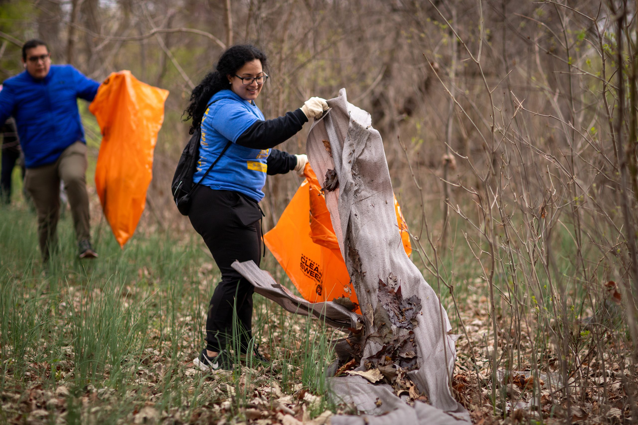 A woman holds a trash bag as she picks up litter along a wooded area.