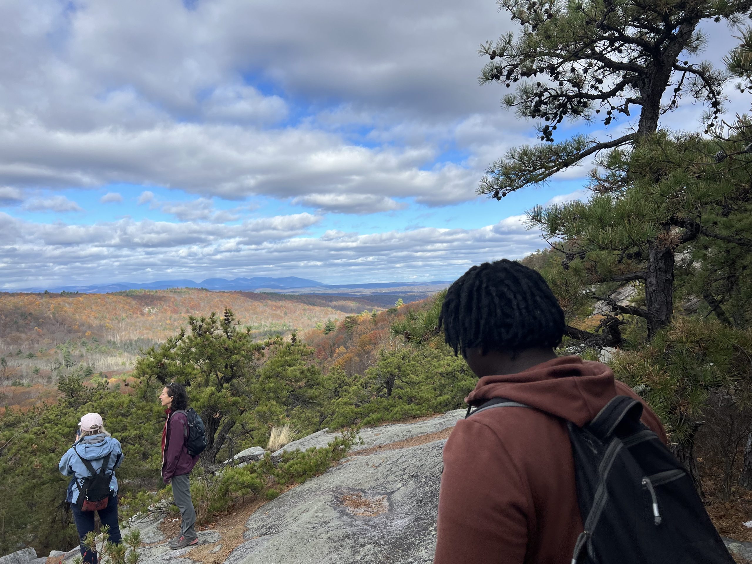 Atop a mountain, three people look toward a mountain range in the fall.