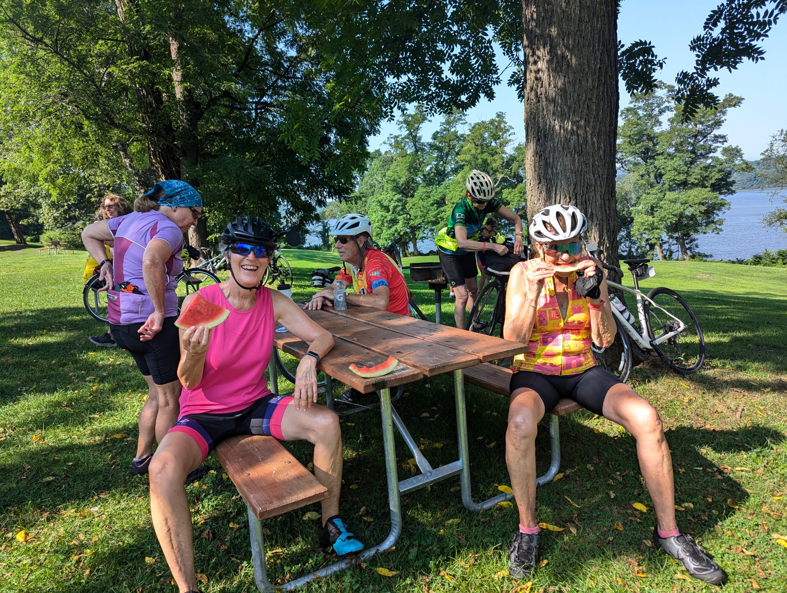 A group of cyclists sit at a picnic table eating watermelon.