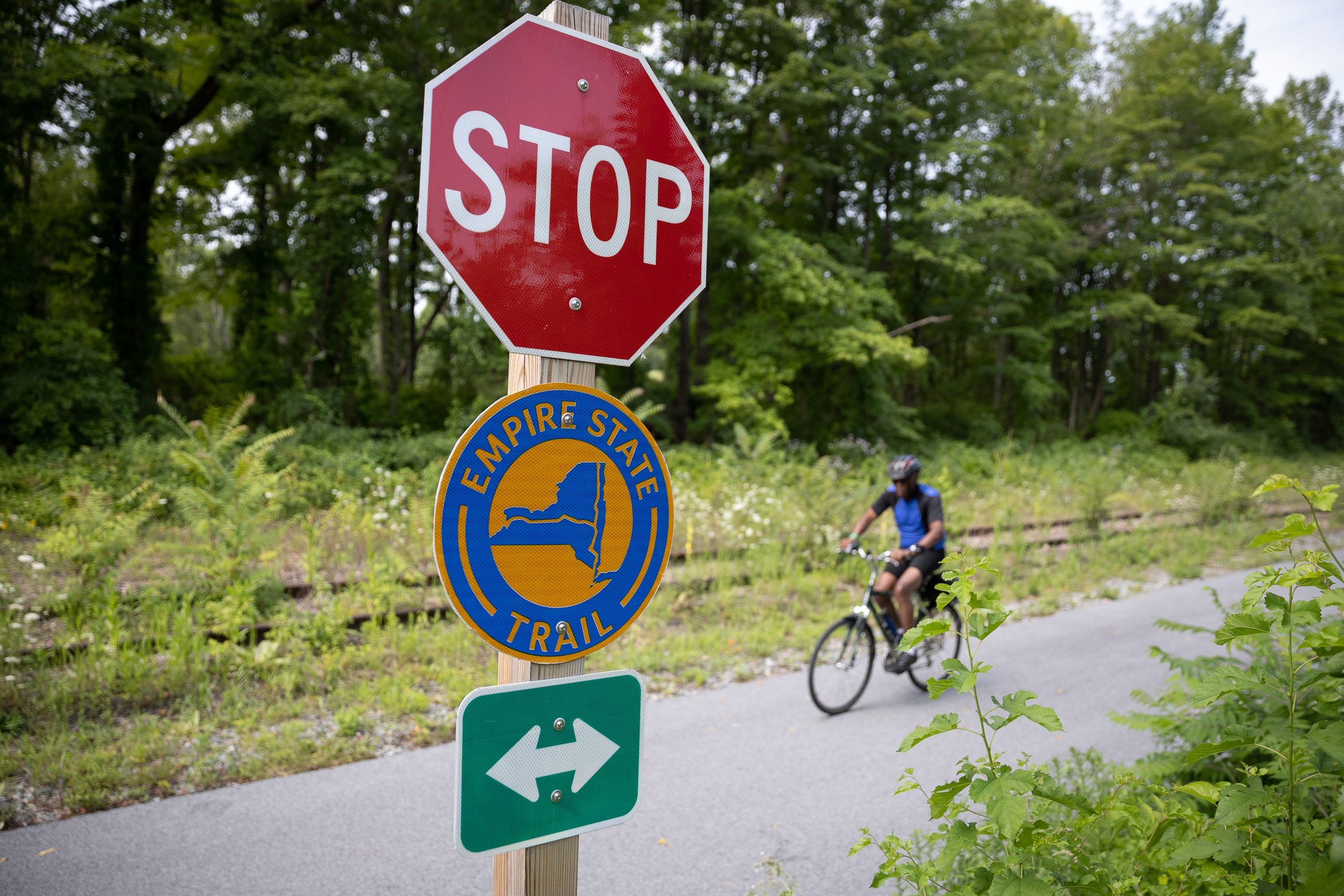 A cyclists rides along a paved path with a Empire State Trail sign in the foreground.
