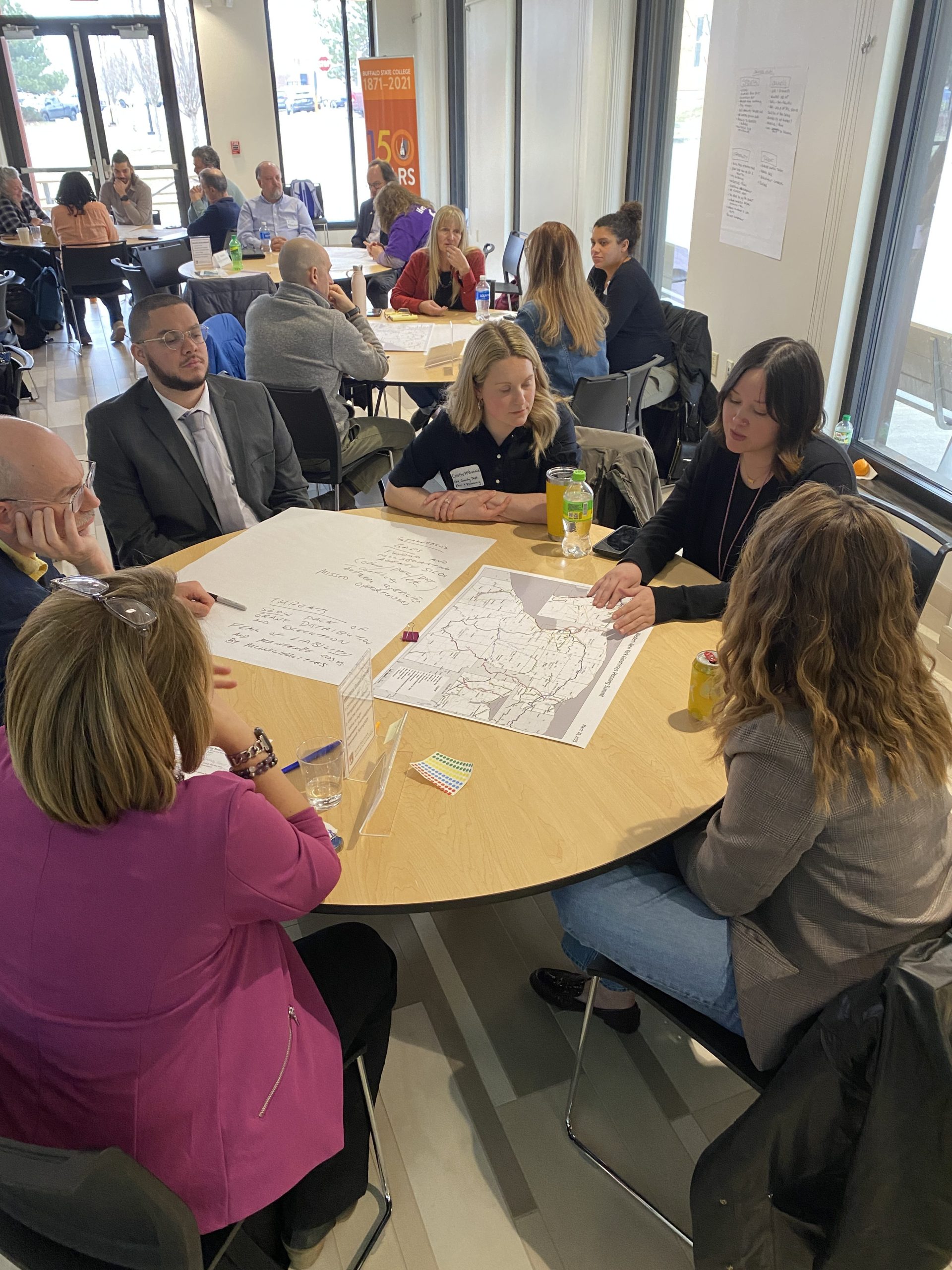 Six people are seated at a table, pointing at and discussing a map of Western New York, with a second large sheet of paper with writing on it summarizing strengths, weaknesses, opportunities and threats. In the background are other tables with other groups of people doing the same activity