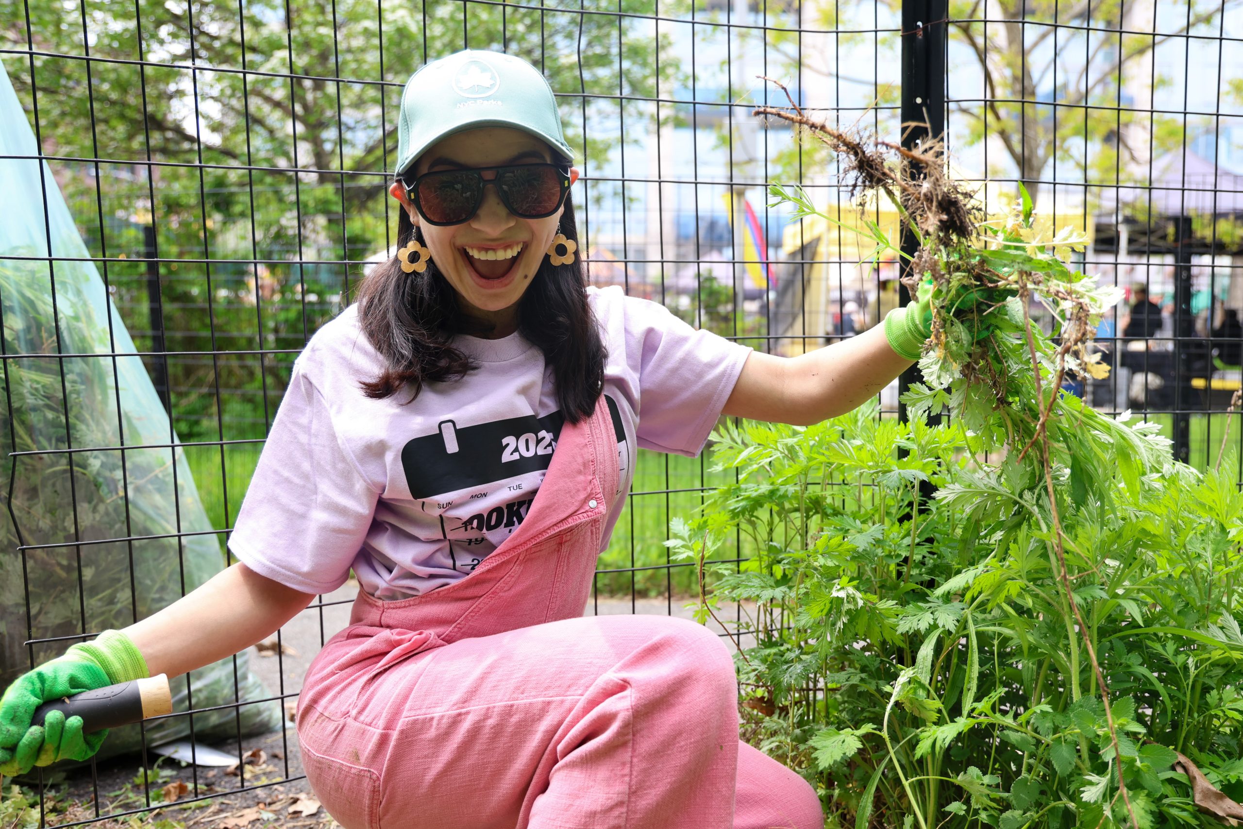 A young woman in pink overalls smiles as she pulls a large weed from a garden at I Love My Park Day.