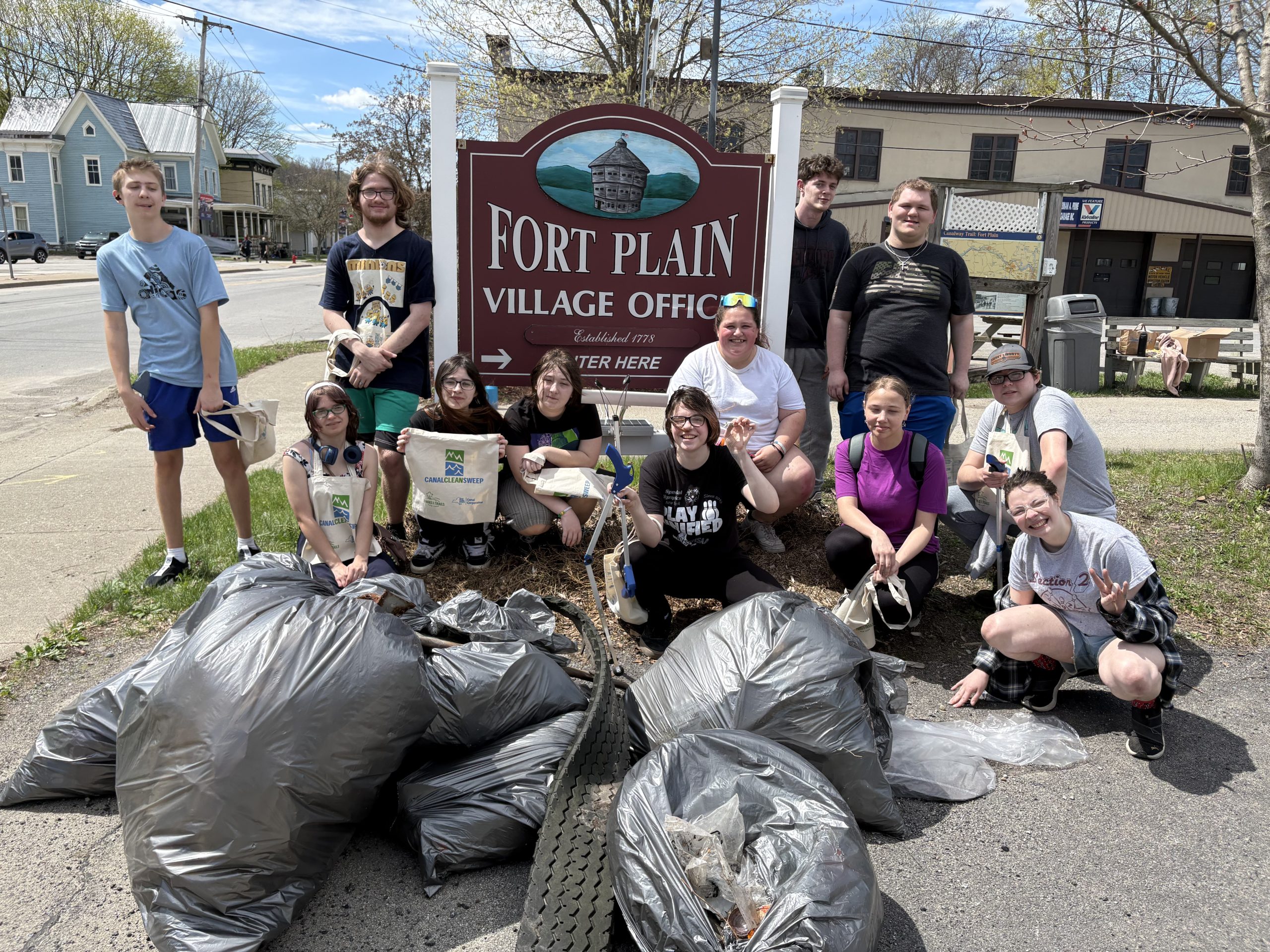 A group of volunteers pose for a photo with full trash bags next to a Fort Plain sign.