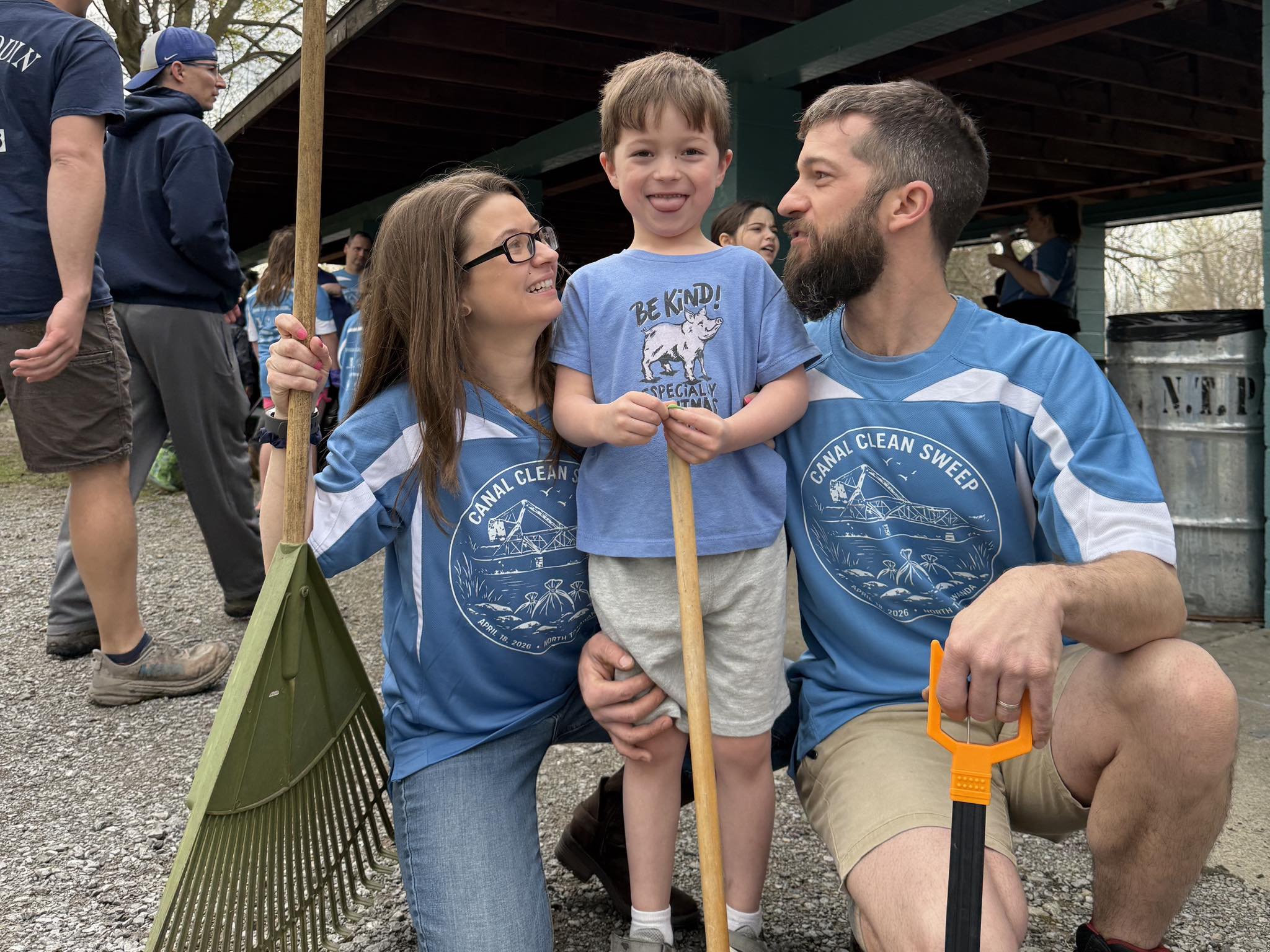 A mother and a father hug their son while holding rakes and other tools.